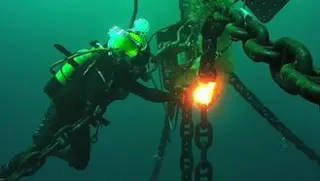 A diver in full gear uses a welding torch to work on a large chain underwater, with bubbles rising to the surface.
