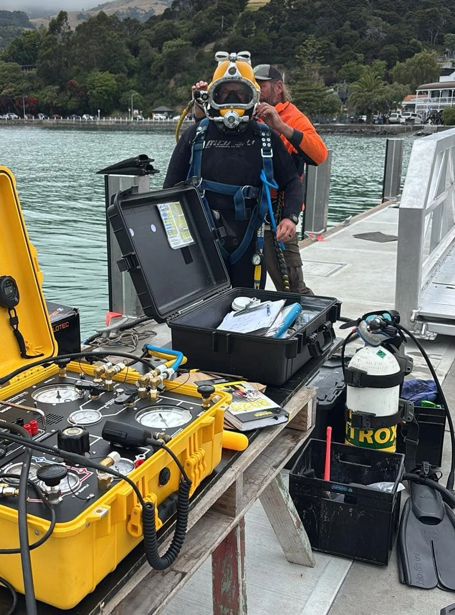 A commercial diver in full gear stands on a dock while another person assists; diving equipment and monitoring devices are arranged on a nearby table in Akaroa