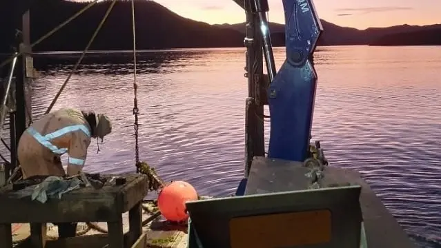 A person in work clothes stands on a boat at sunset, inspecting equipment near the water with mountains visible in the distance.