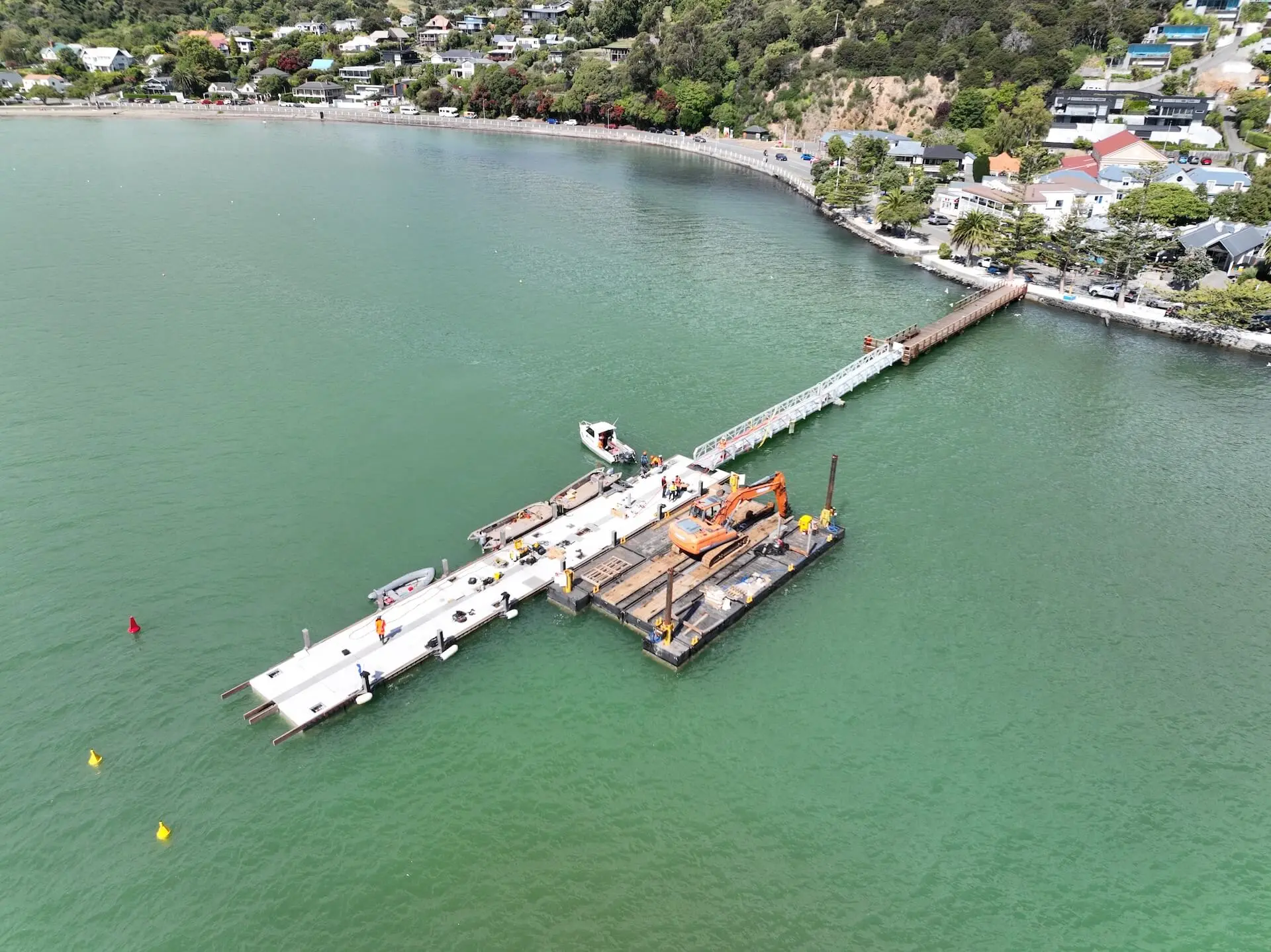 Aerial view of a construction barge with cranes and workers building a pier extending from the shoreline into the water near a coastal town.