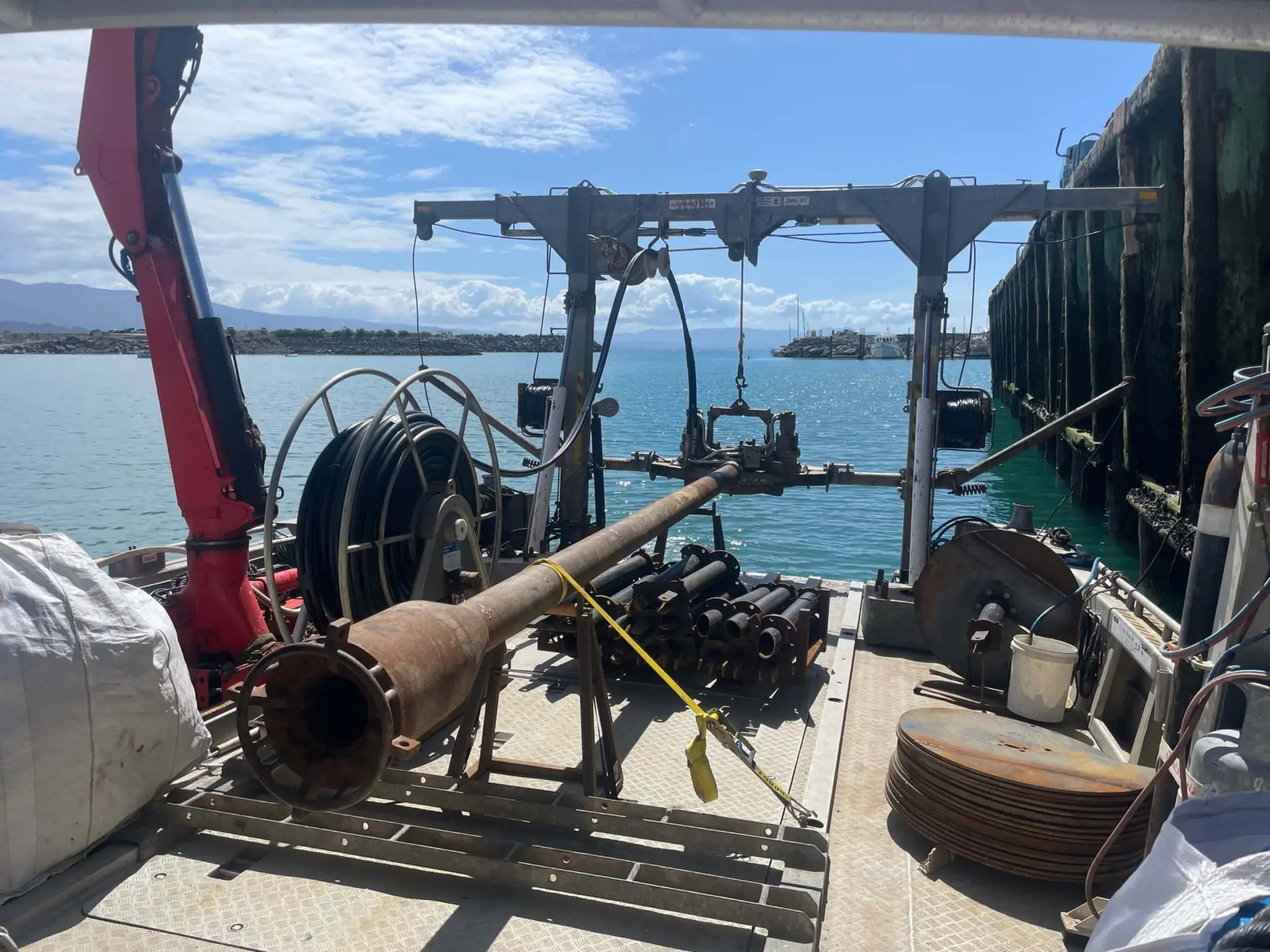 A metal work platform on a docked boat holds large pipes, cables, and machinery with a water and sky backdrop.