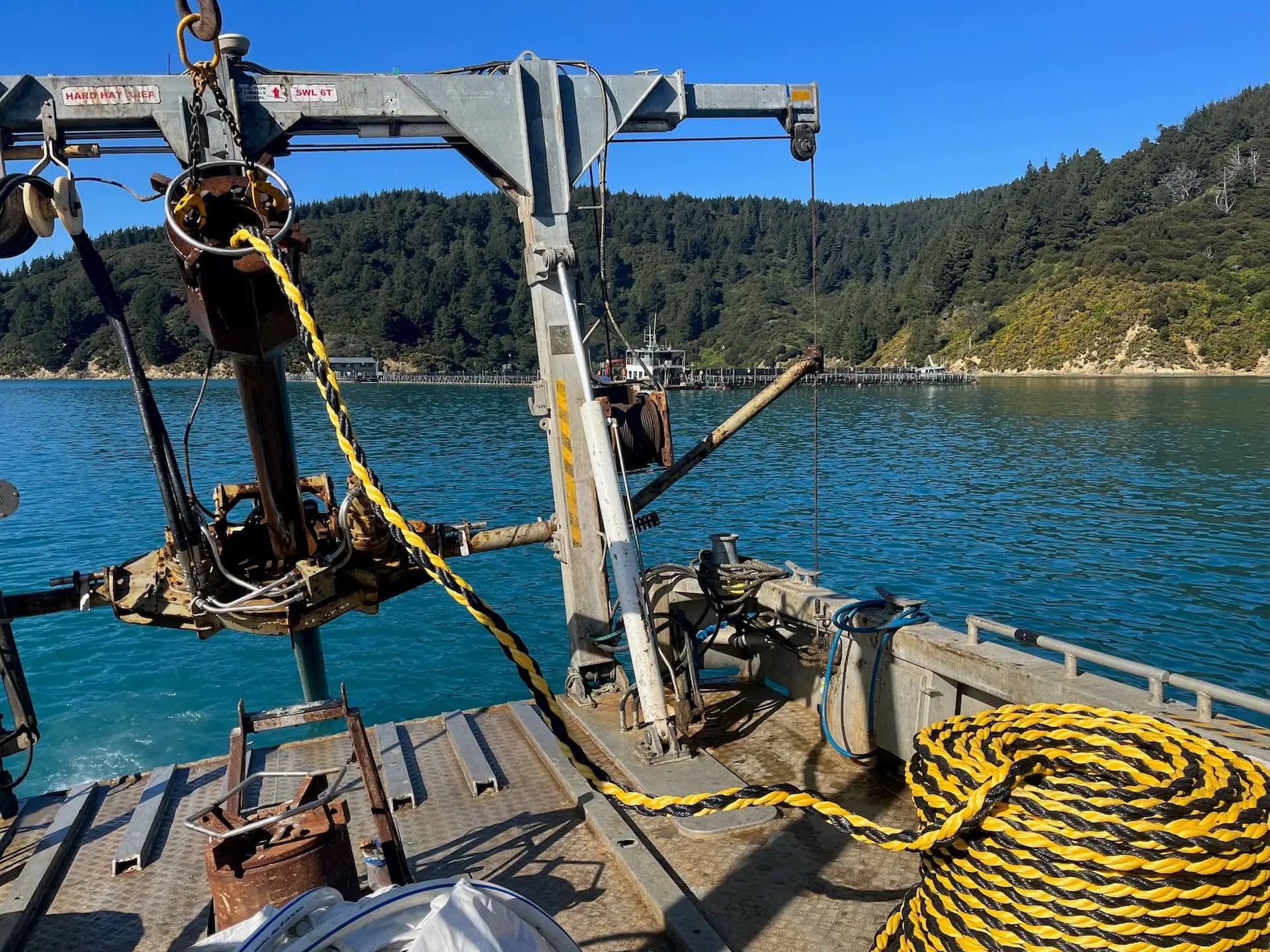 A boat deck outfitted with marine equipment is pictured on the water near a forested shoreline under a clear blue sky.