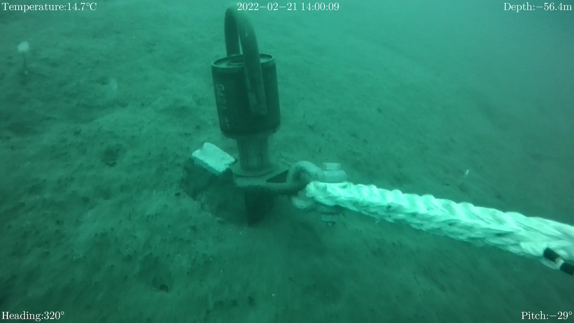 Underwater view of a large anchor shackle attached to a rope on the seabed, with temperature, date, depth, heading, and pitch displayed on the screen.