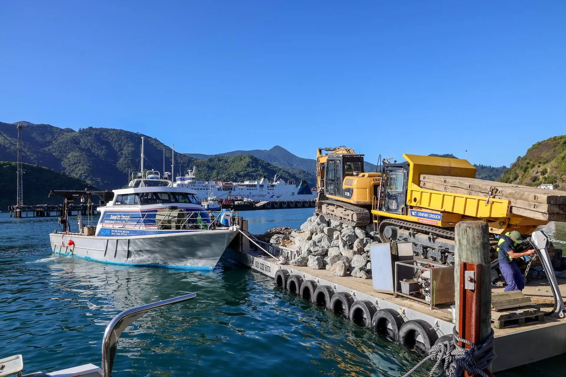A barge carrying rocks and construction equipment is docked next to a work boat in a harbor with mountains in the background.