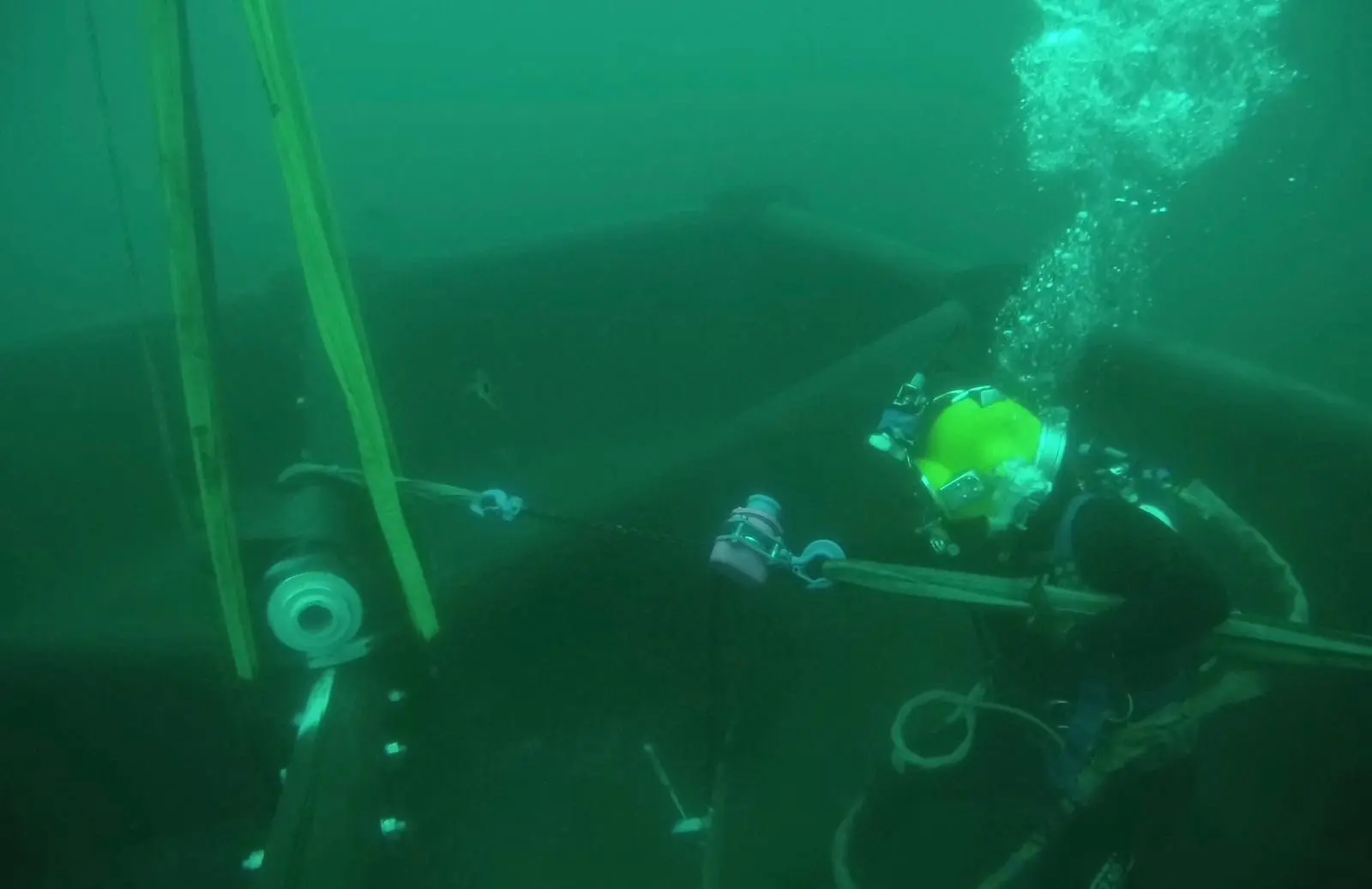 A diver wearing scuba gear works underwater, holding a tool near large industrial equipment while air bubbles rise to the surface.