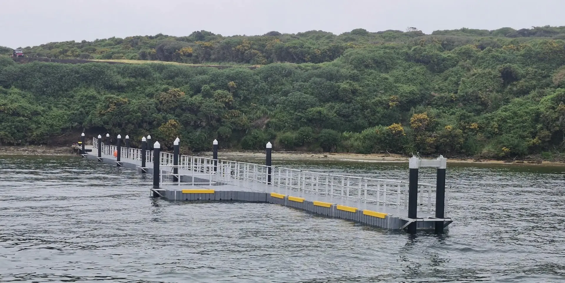 A floating jetty with railings and yellow markings extends over calm water toward a forested shoreline under an overcast sky.