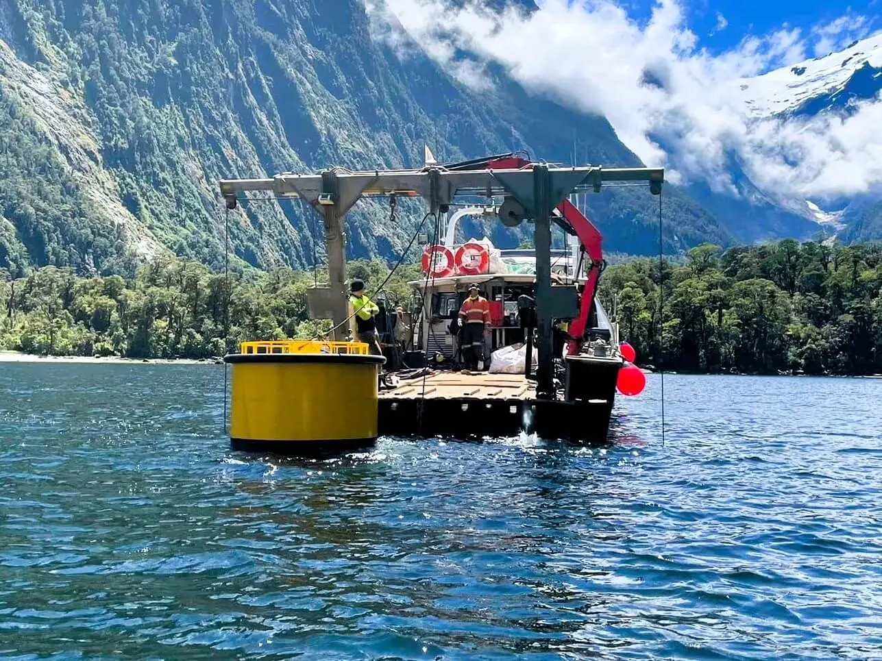 A vessel with equipment and red buoys is positioned on a lake near a mountainous forest shoreline under a partly cloudy sky.