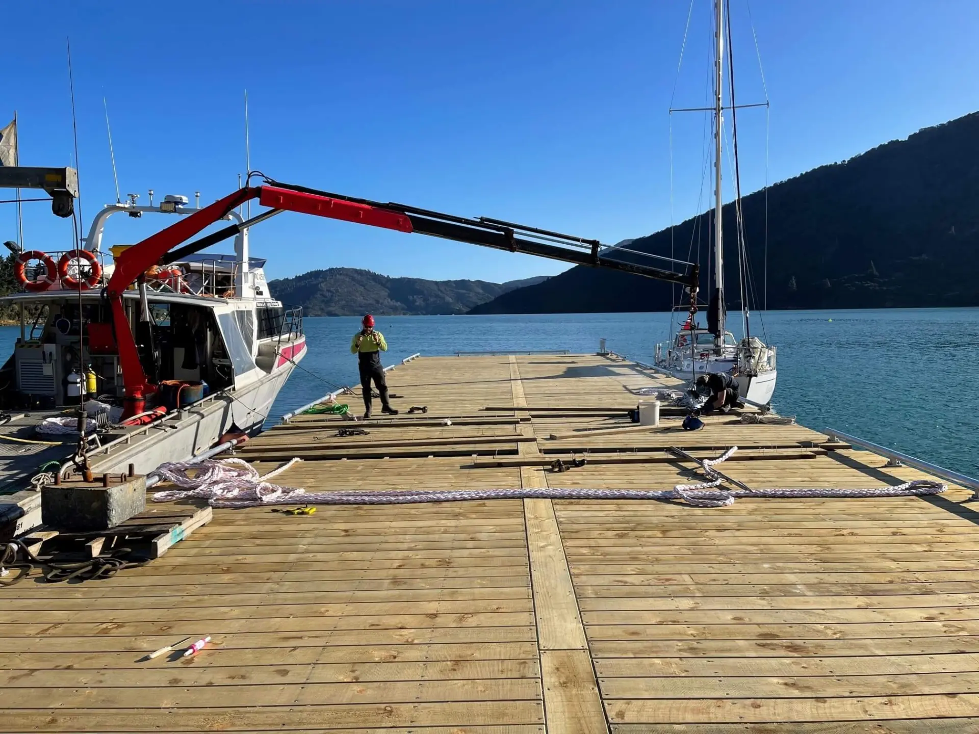 A person stands on a wooden dock next to two boats, with a crane arm extended over the dock. Mountains and water are visible in the background under a clear sky.
