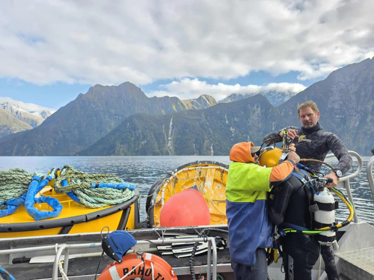 Two people on a boat prepare scuba diving gear, with yellow equipment and rope nearby, against a backdrop of mountains and water under a partly cloudy sky.