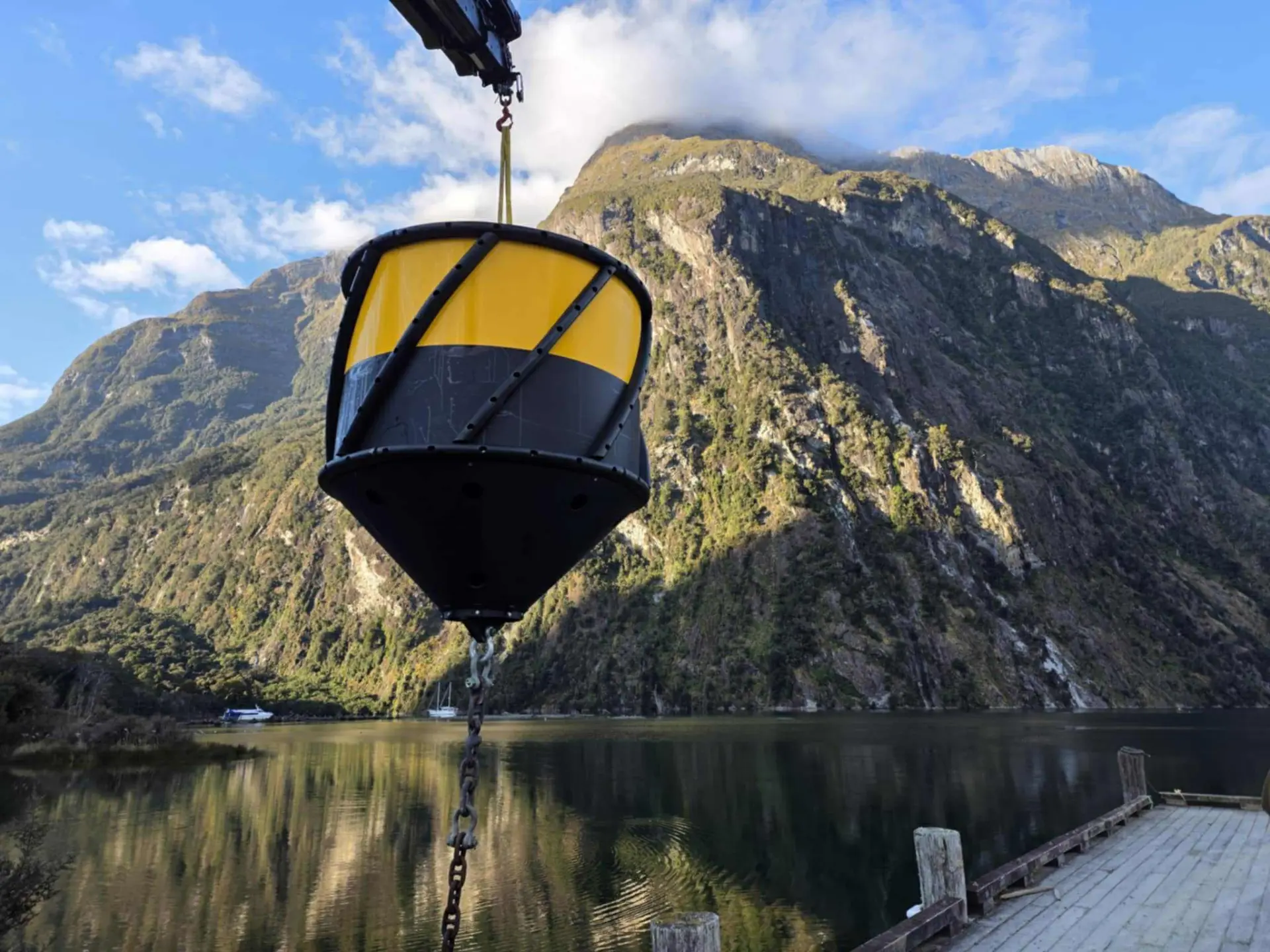 A large yellow and black buoy is suspended by a crane over a calm lake, with steep mountains and clear sky in the background.