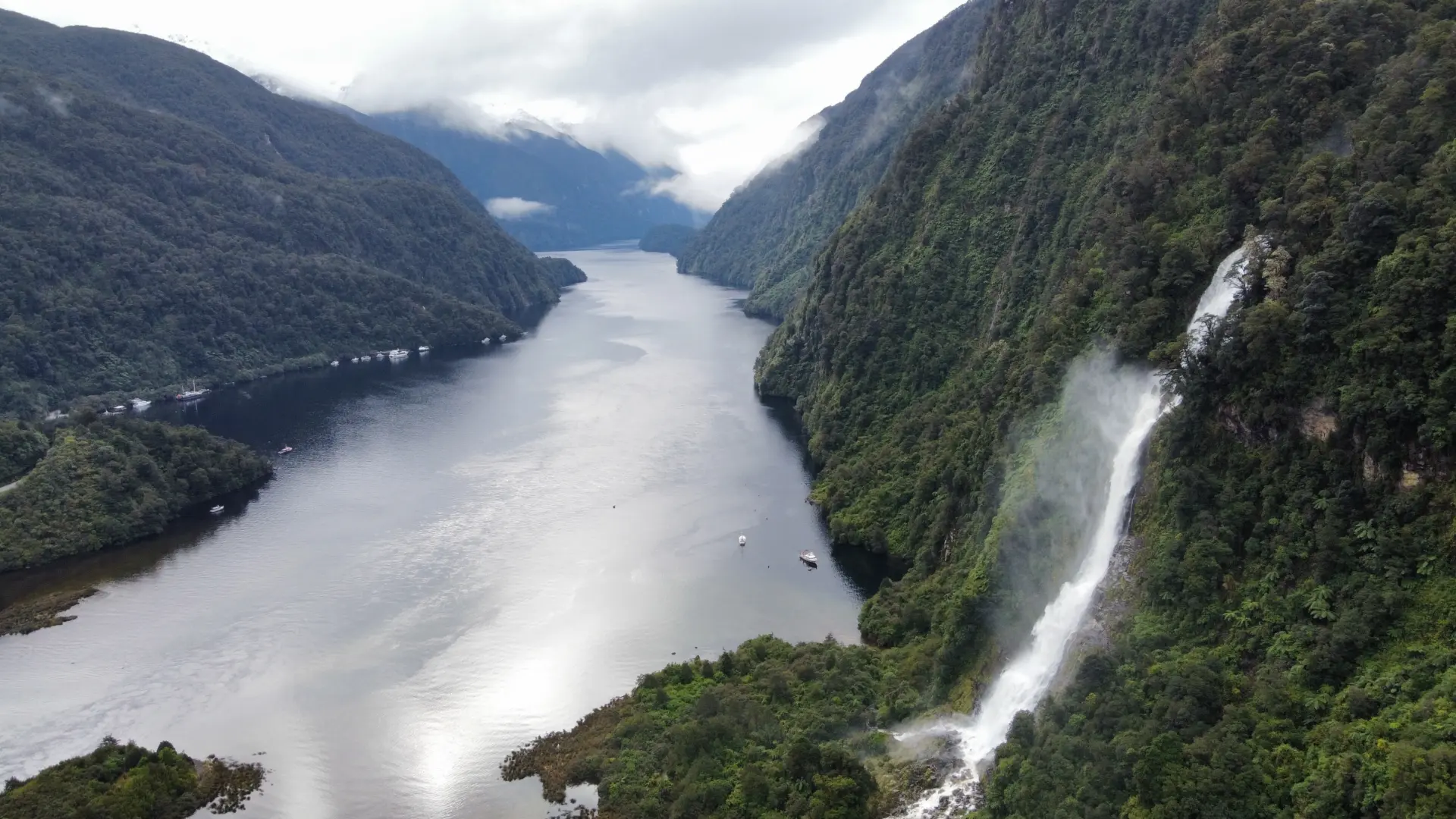 Aerial view of a river running between steep, forested mountains with a waterfall cascading down the right slope under a cloudy sky.