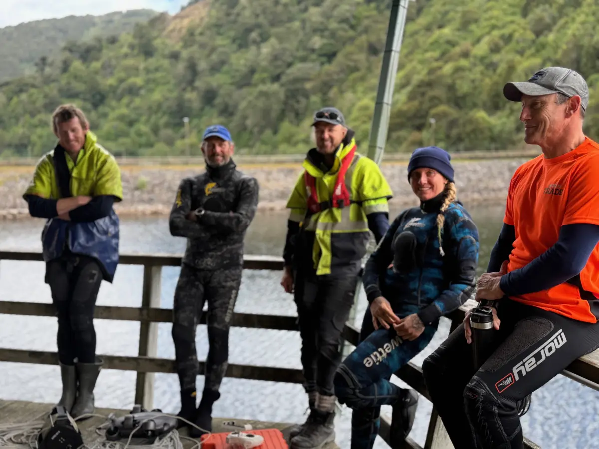 Five people in wetsuits and outdoor gear stand and sit on a dock by the water, with trees and hills visible in the background.