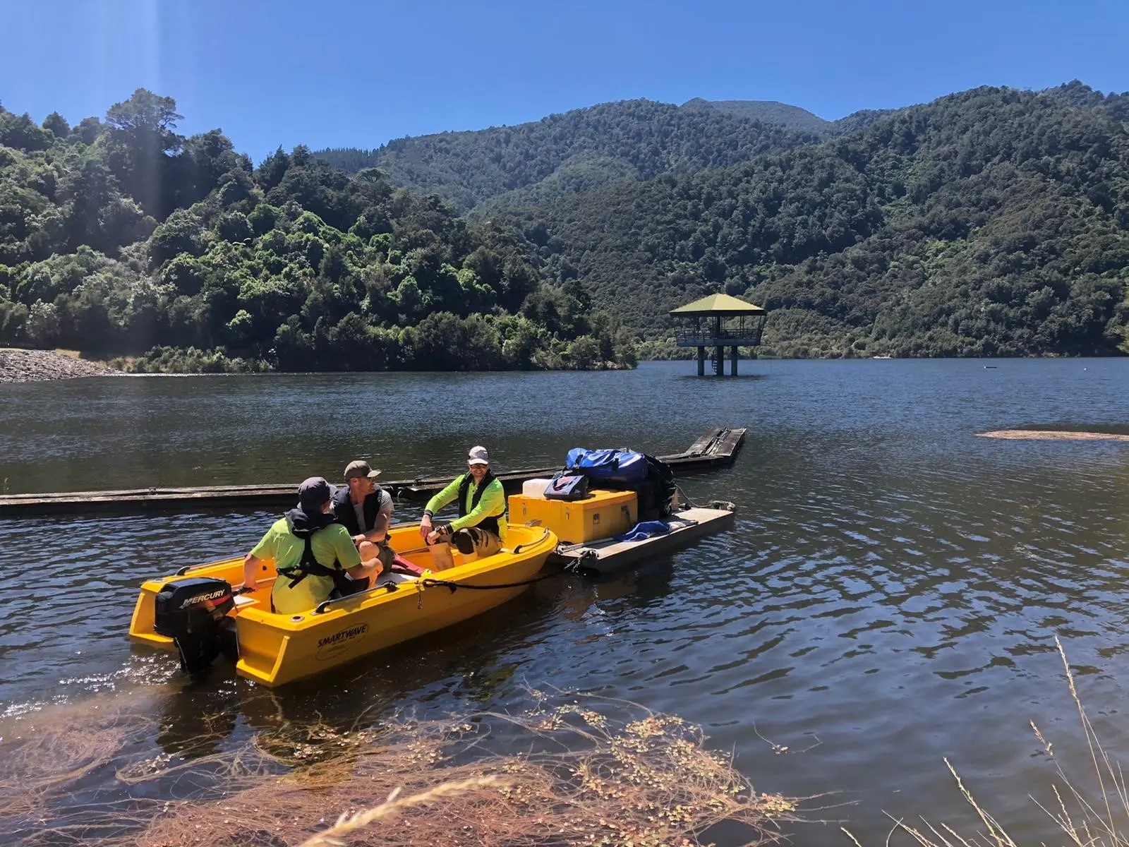 Three people in yellow motorized boats with equipment on a lake, surrounded by forested hills and mountains under a clear blue sky.