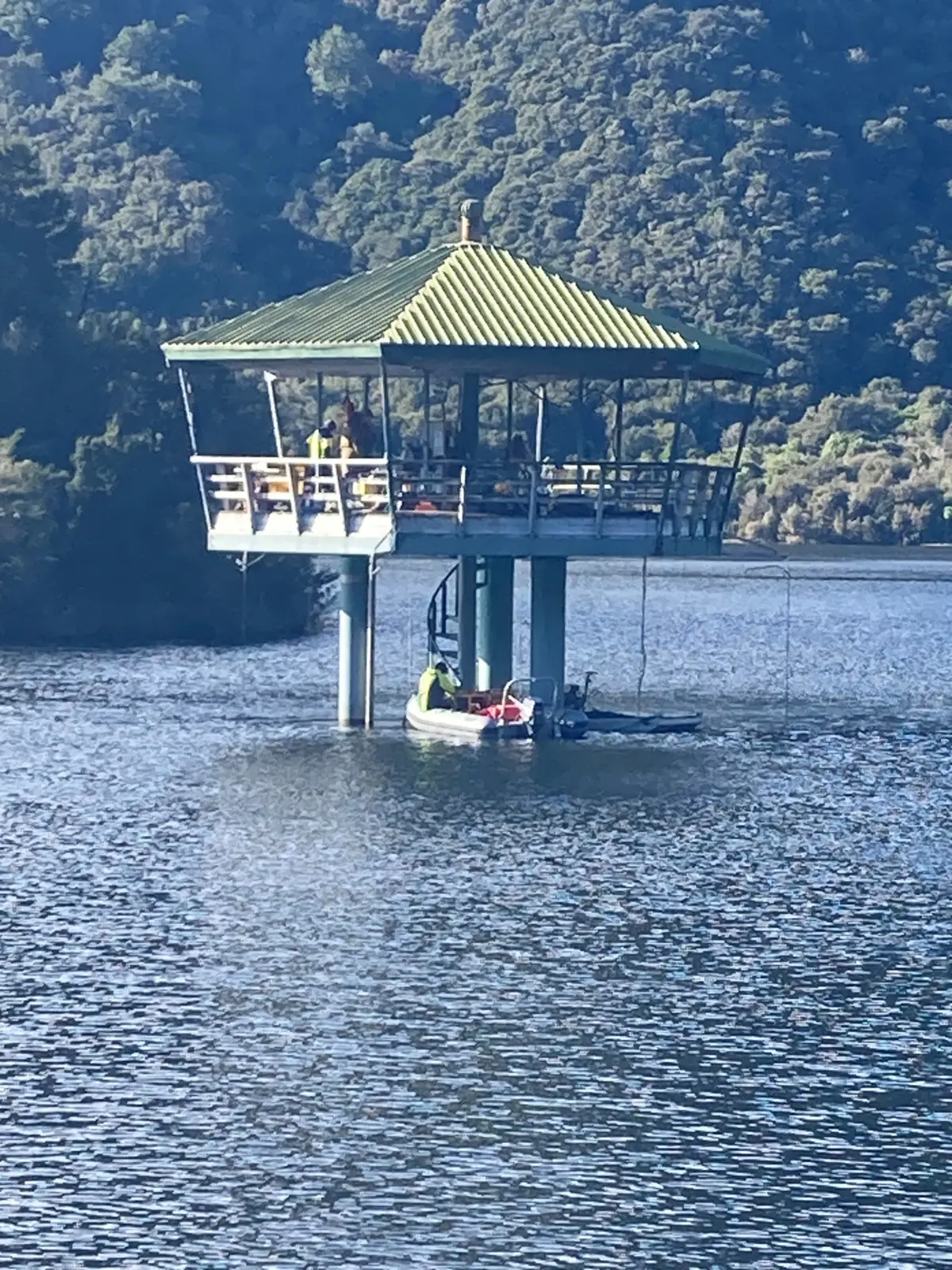 A green-roofed structure on stilts stands over a lake, with a small boat docked beneath it and wooded hills in the background.