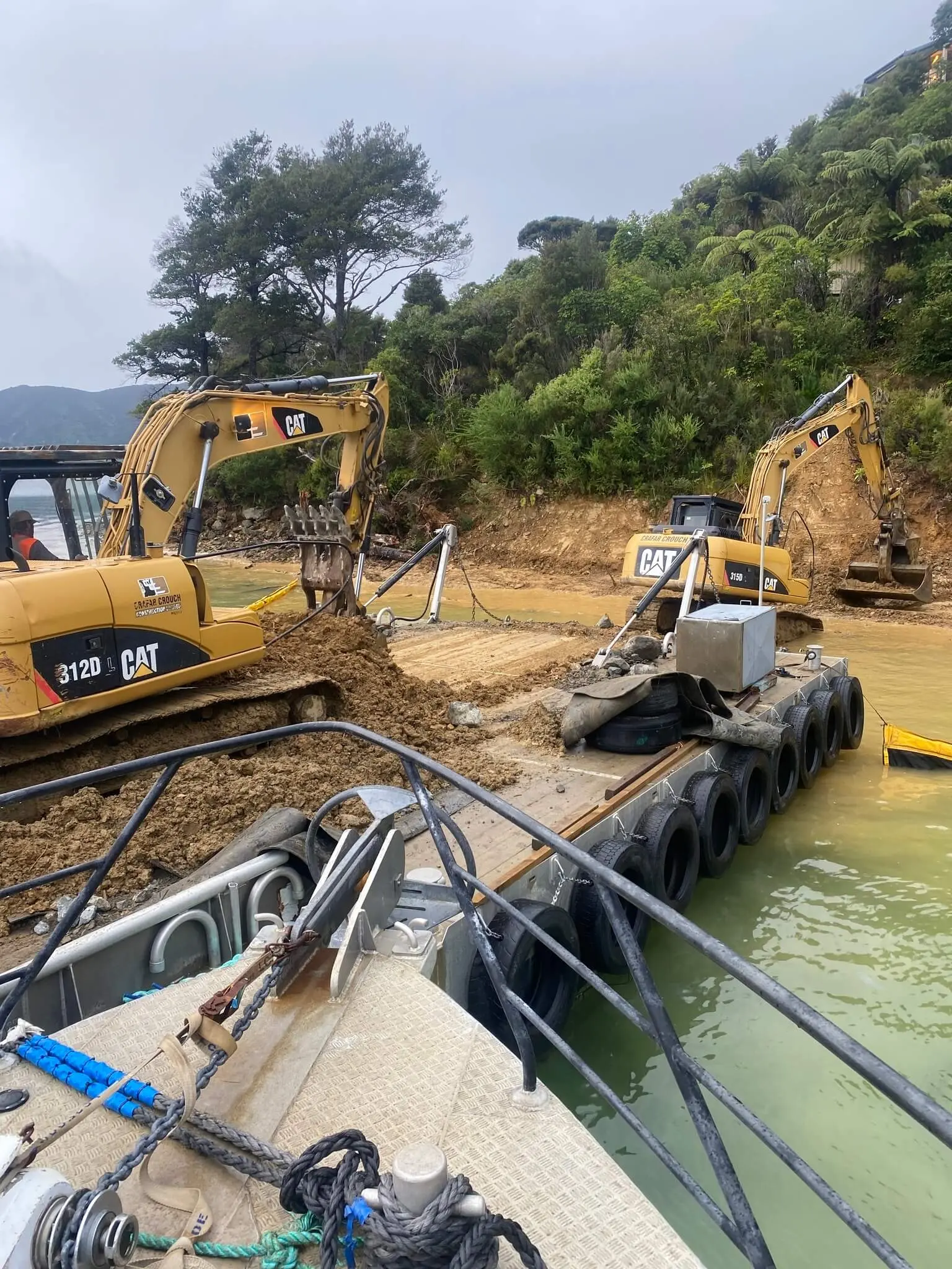 Two excavators work on the shoreline, moving earth near a floating barge on a greenish body of water, with forested hills in the background.