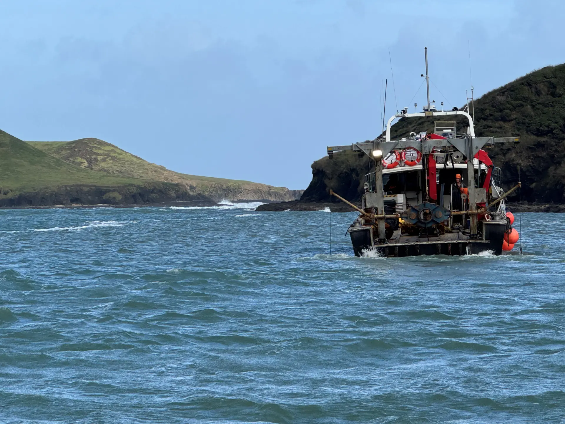 A fishing boat sails away from shore on a blue ocean, with green hills and rocky cliffs in the background under a clear sky.